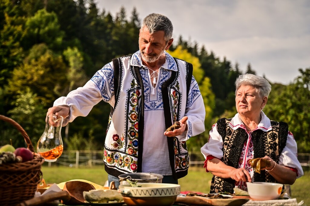 Un bărbat și o femeie în port popular, la o masă pe iarbă, toarnă dintr-o sticlă